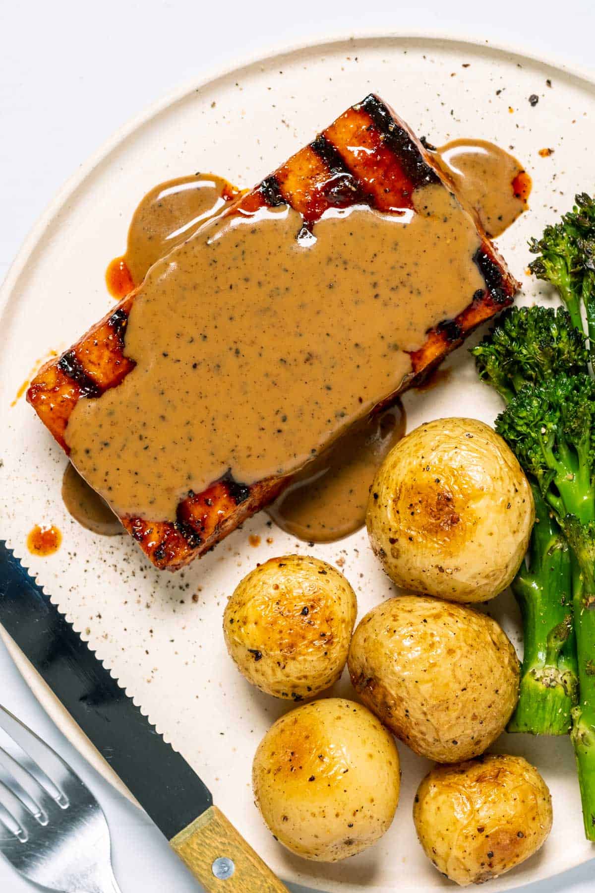 Tofu steak topped with peppercorn sauce on a white plate with baby potatoes and broccoli.