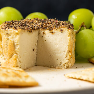 Vegan camembert with slices removed on a white plate with crackers and grapes.