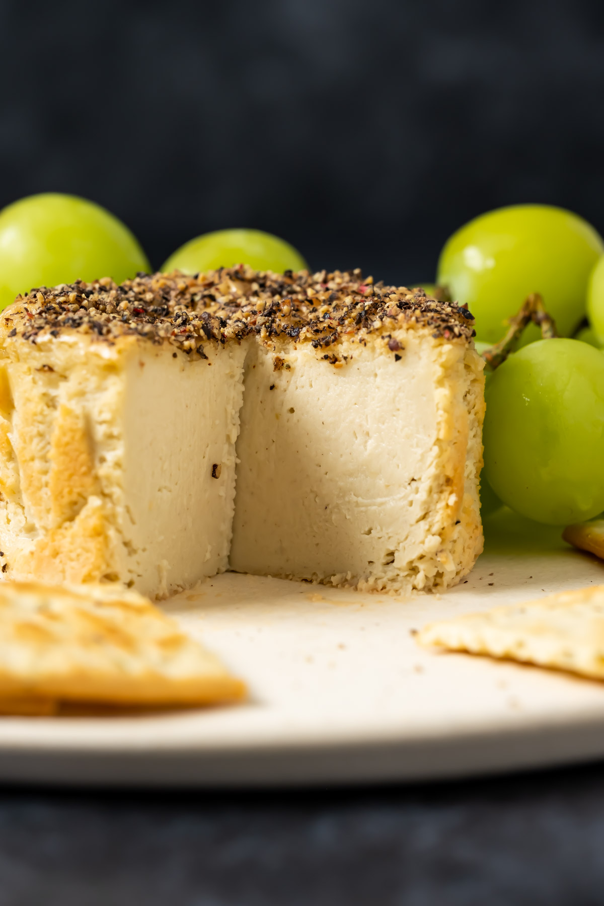 Vegan camembert with slices removed on a white plate with crackers and grapes.