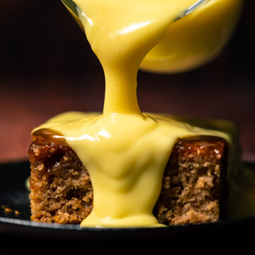 Custard pouring from a glass jug over a slice of sticky toffee pudding.