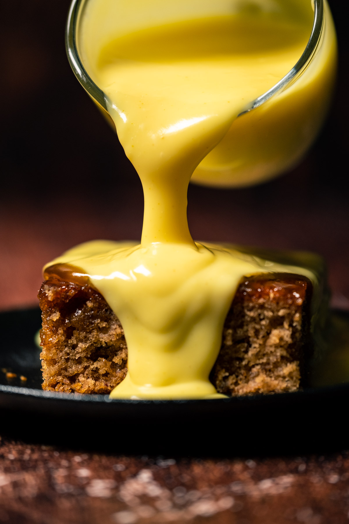 Custard pouring from a glass jug over a slice of sticky toffee pudding.