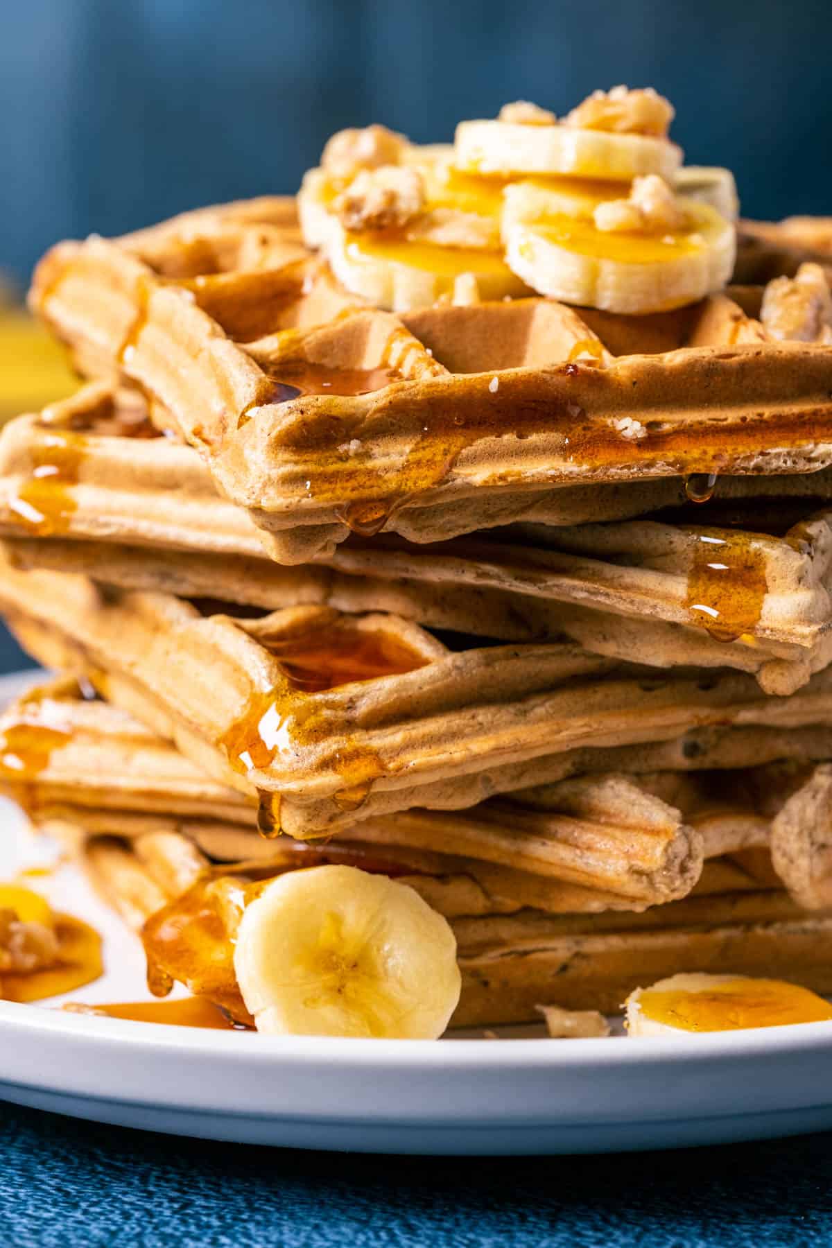 Stack of vegan banana waffles topped with sliced banana, walnuts and syrup on a white plate.