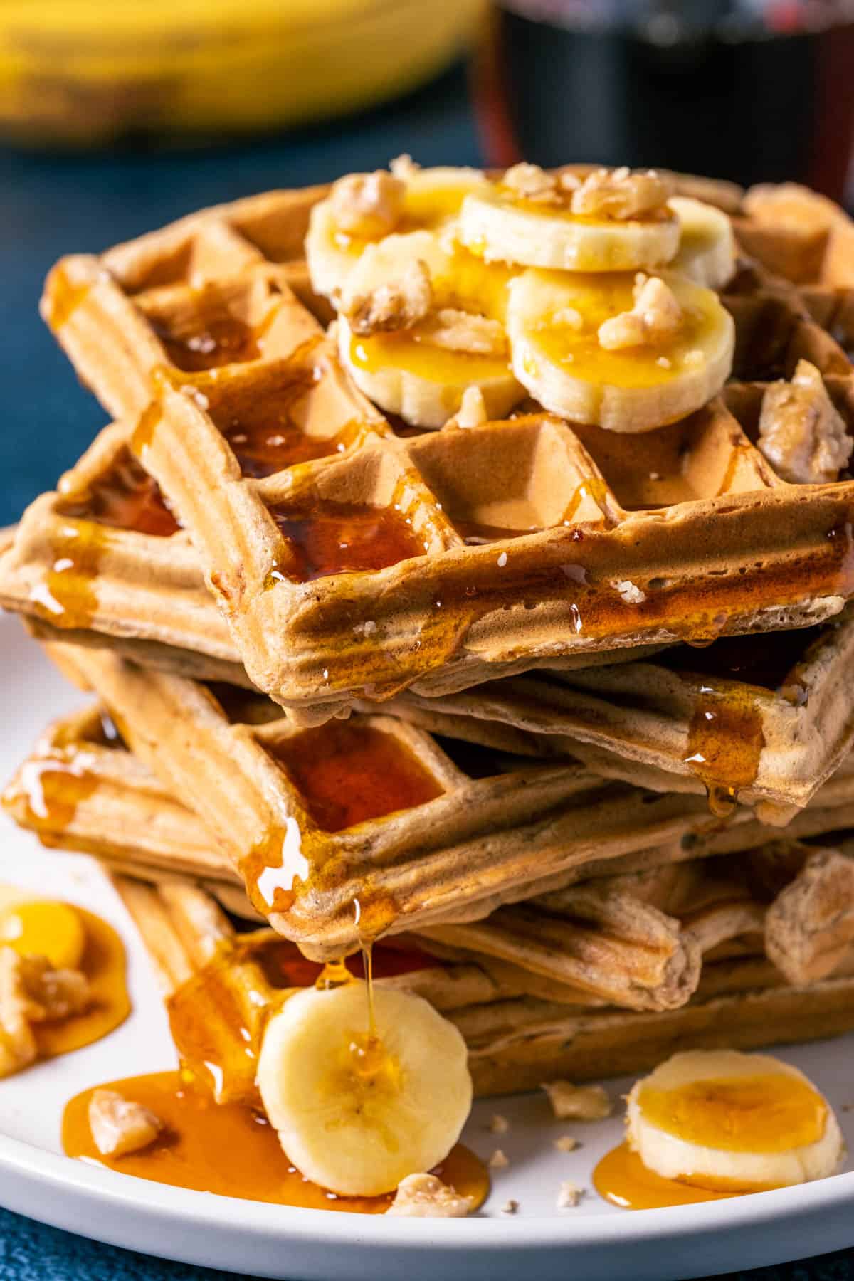 Stack of vegan banana waffles topped with sliced banana, walnuts and syrup on a white plate. 