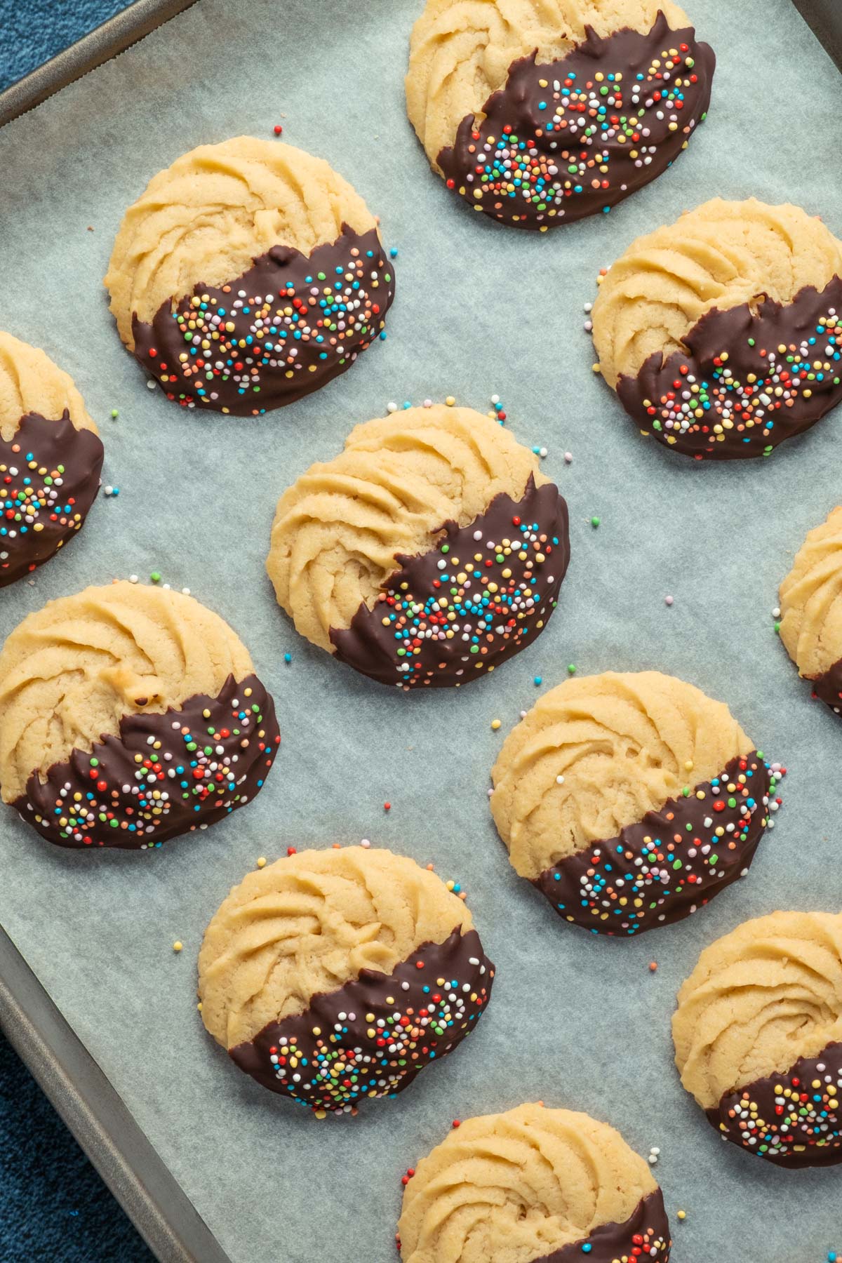 Butter cookies dipped in chocolate and topped with sprinkles on a parchment lined baking tray. 