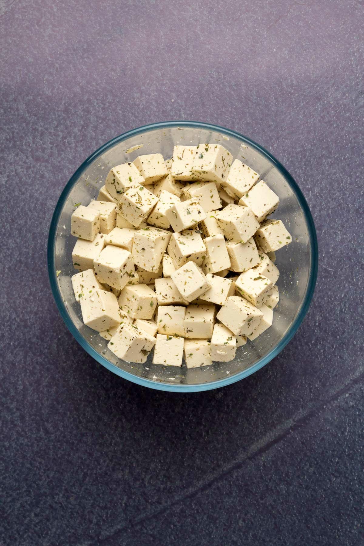 Cubes of vegan feta in glass bowl with dried herbs.